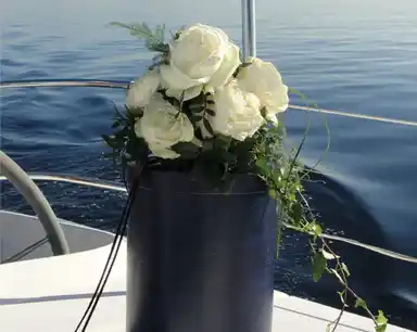 Eine Urne mit Blumenstrauß steht auf einem Schiffsdeck, im Hintergrund ist das Meer