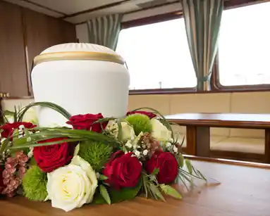 Eine Urne mit Blumenstrauß steht auf einem Tisch in einer Schiffskajüte, im Hintergrund sind Fenster